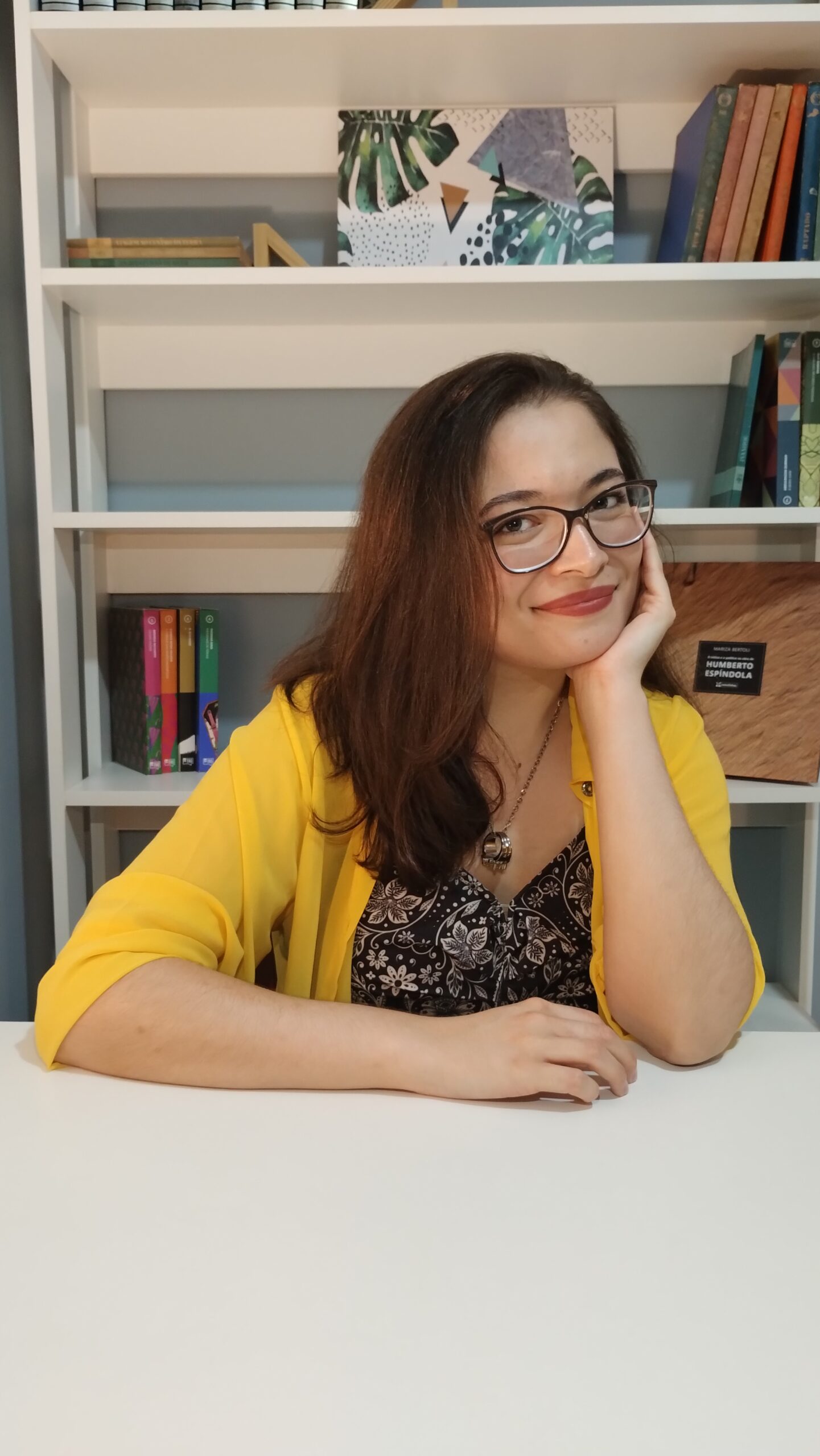 Foto da Psicóloga Roberta Damasceno. Mulher de cabelos castanho-claros, pele branca, usando óculos no rosto e sorrindo. Veste uma blusa amarela de mangas longas sobre uma vestimenta escura com estampas claras. Está apoiando o rosto na mão. Atrás dela tem uma estante com livros coloridos.